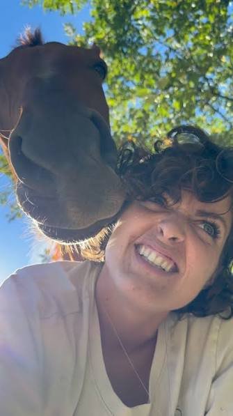 A chestnut horse nuzzling the head of a smiling woman under green trees